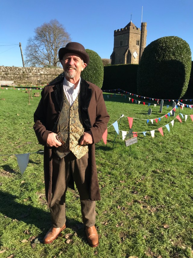 Wel standing on a green lawn, dressed in brown Victorian men's costume with a church in the background